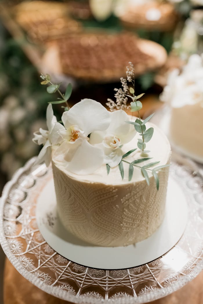 about-05 A beautifully decorated wedding cake adorned with white flowers and leaves on a glass stand.